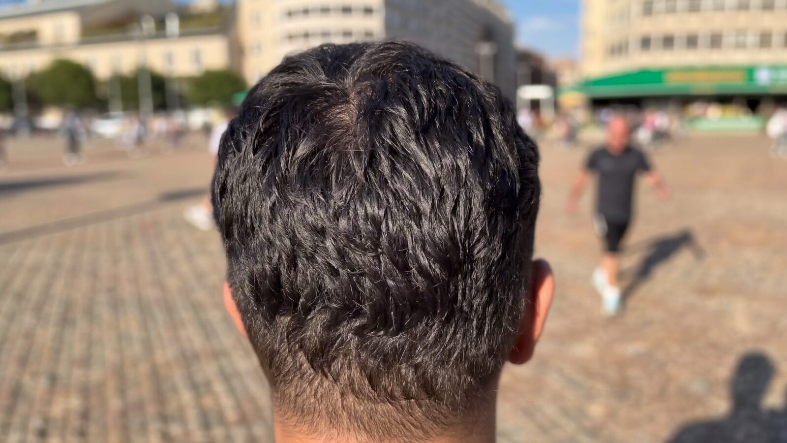 Man seen from behind with full, healthy hair on the crown in a sunny plaza
