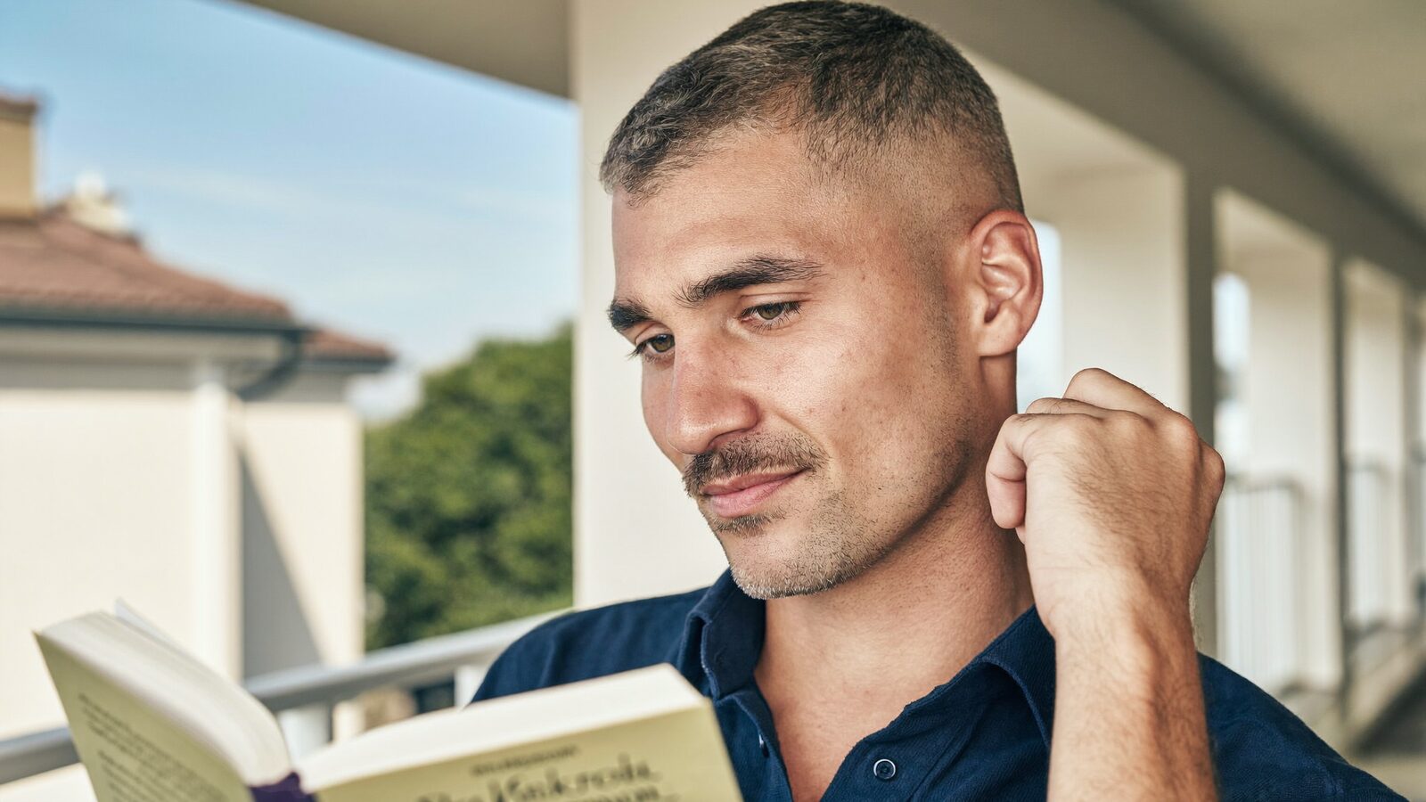 Man with short hair reading a book on a sunny balcony