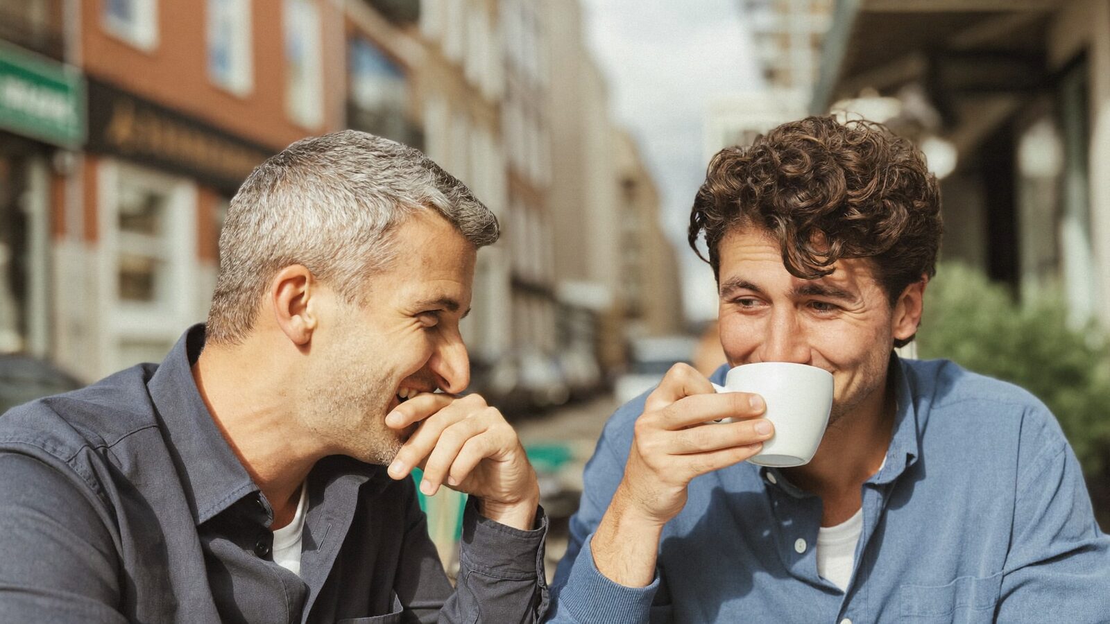 Two friends with full hair laughing over coffee at a sidewalk cafe