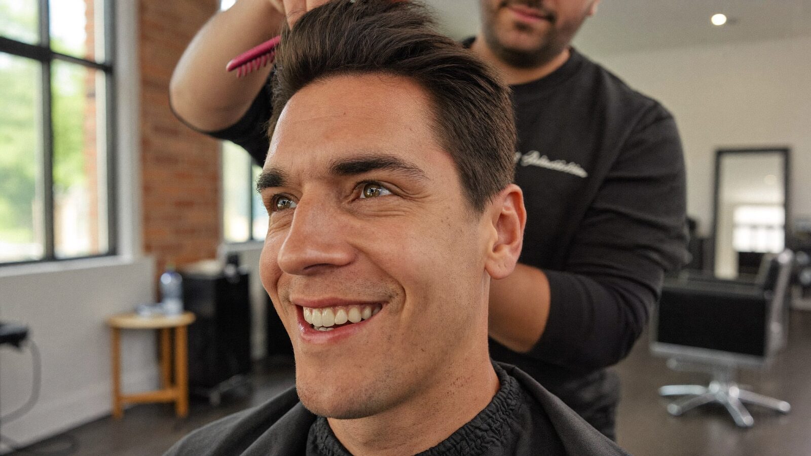 Smiling man with short dark hair in a salon chair while a stylist combs his hair