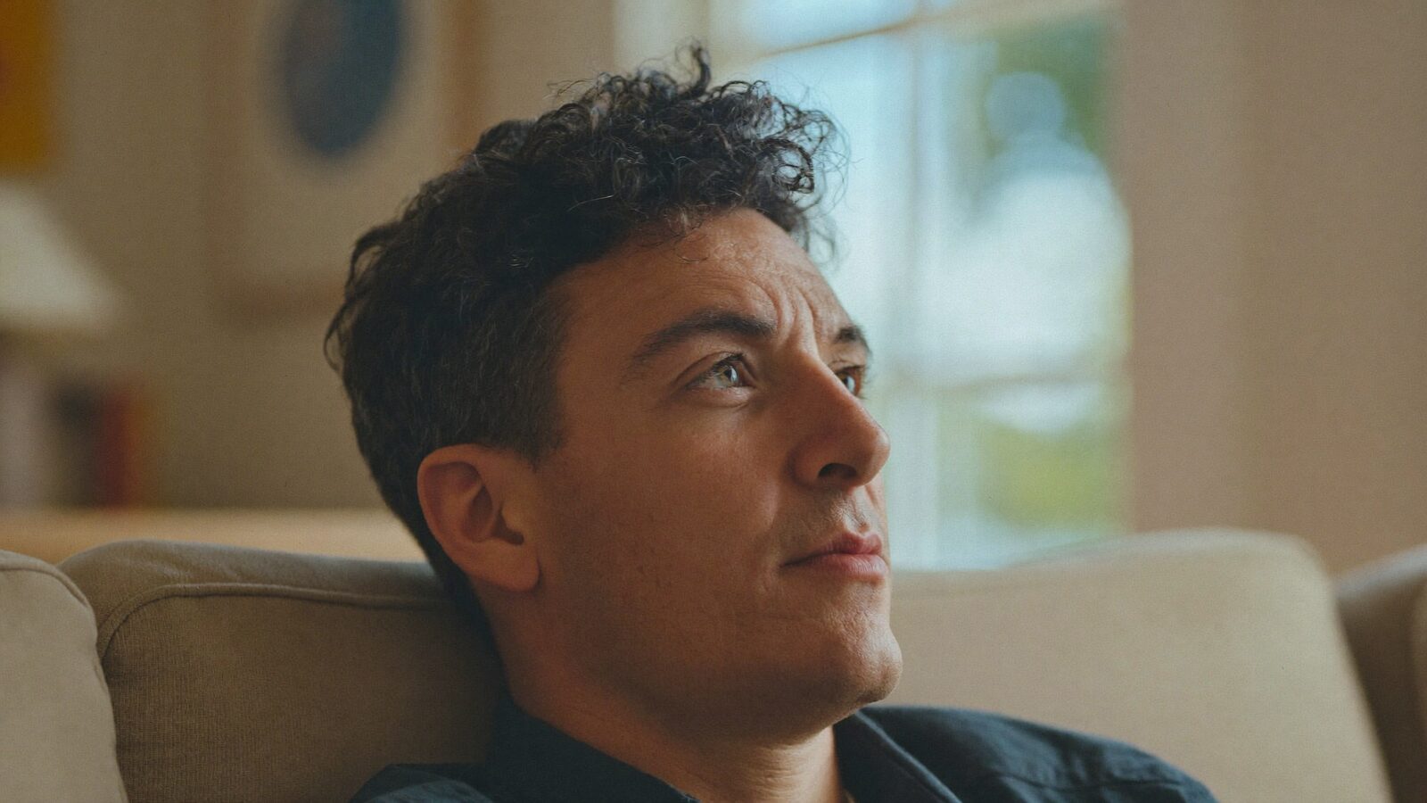 Man with full, curly dark hair relaxing on a sofa in a bright room