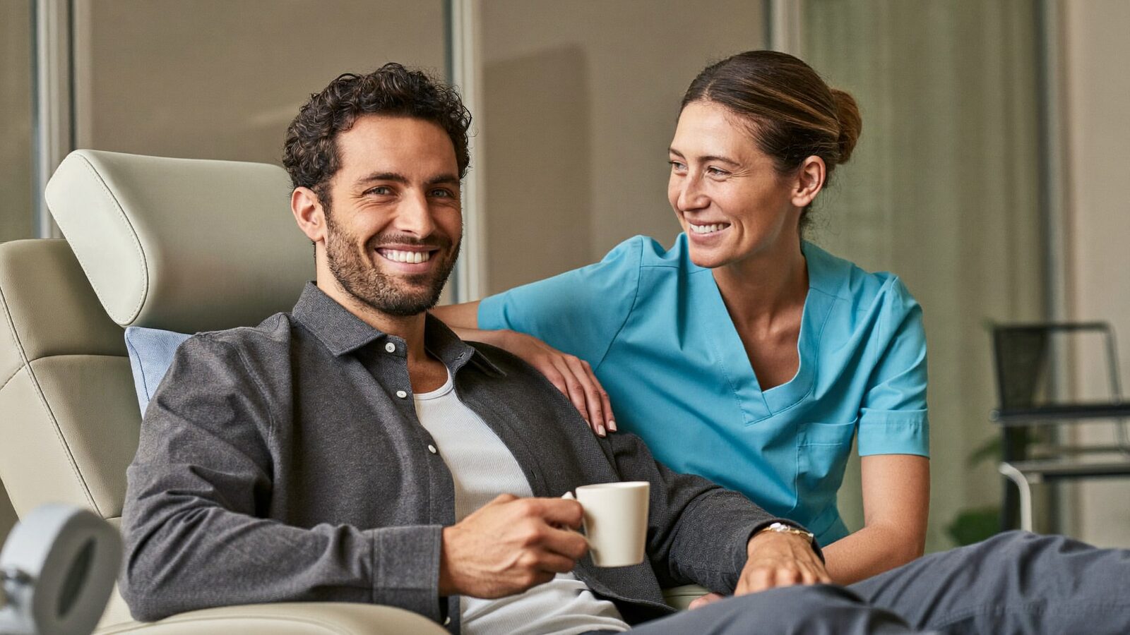 Smiling man with a cup of tea in a recovery chair beside a nurse