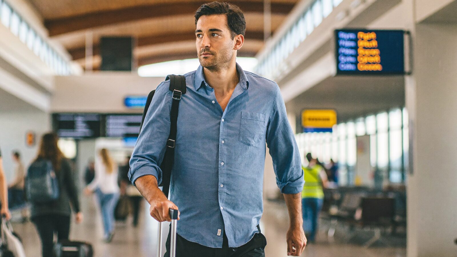 Man with full hair walking through an airport terminal with a suitcase