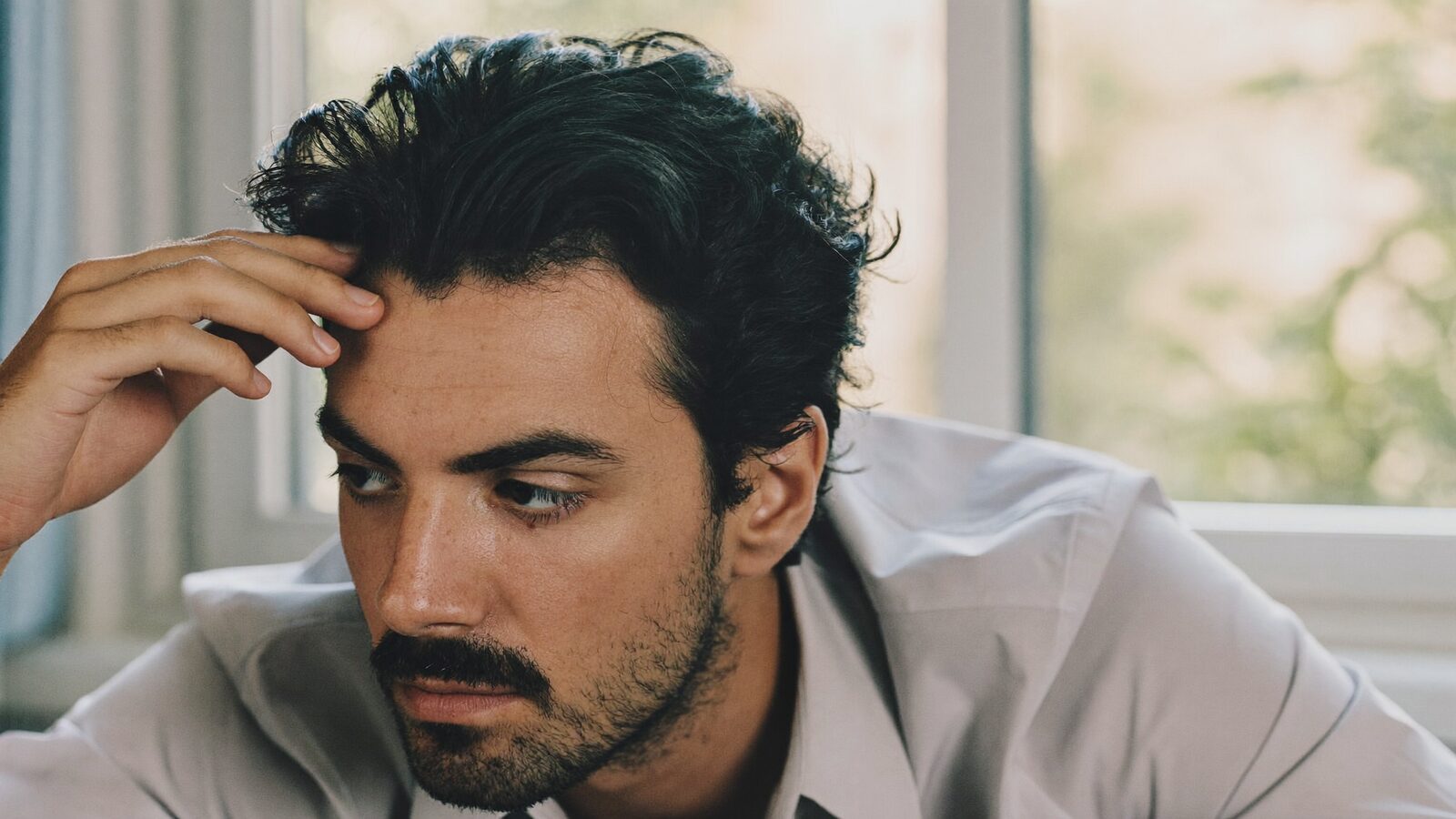 Man with thick dark hair touching his hairline near a bright window